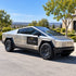 A silver Tesla Cybertruck-style pickup truck parked outdoors on a paved road, with trees, hills, and a building visible in the background. A black door patch reads 'City One Property' in gold text.