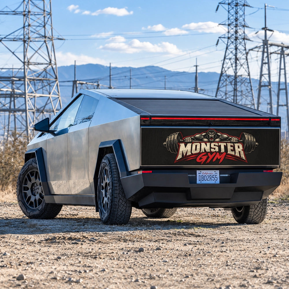 A silver Cybertruck-style vehicle parked outdoors on a gravel lot near power lines and mountains, shown from the rear three-quarter angle. The back panel has a large black graphic reading 'Monster Gym' with red and gold details.