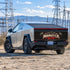A silver Cybertruck-style vehicle parked outdoors on a gravel lot near power lines and mountains, shown from the rear three-quarter angle. The back panel has a large black graphic reading 'Monster Gym' with red and gold details.