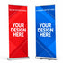 Two retractable banner stands shown on a white studio background: a red deluxe stand on the left and a blue standard stand on the right, each with large placeholder text reading 'YOUR DESIGN HERE' and smaller text indicating deluxe/standard retractable.