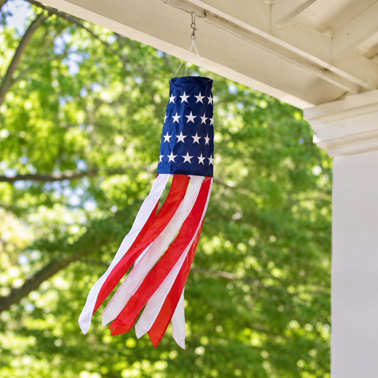 An American flag wind windsock hanging from a porch beam, with a blue top section featuring white stars and red and white flowing ribbon tails, set against a blurred background of green trees.