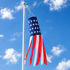 An American flag windsock with a long red and white hanging tails, attached to a silver flagpole against a bright blue sky with scattered white clouds.