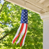 An American flag-themed wind spinner hanging from a porch ceiling, with a blue top section featuring white stars and red and white ribbon-like tails, set against a blurred background of green trees and sunlight.